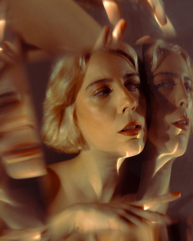Photographic portrait fragmented through mirrored planes: a woman's face repeats and refracts in warm amber light, splitting into overlapping reflections.