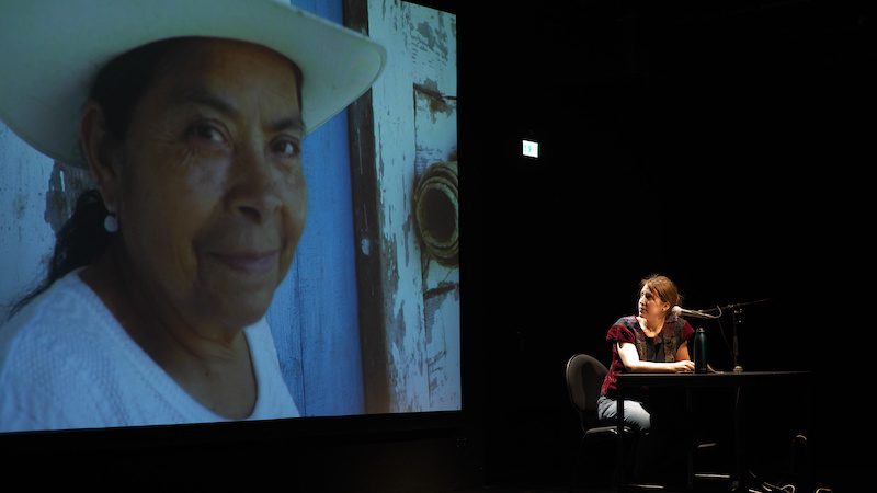 A performer sits at a table with a microphone on a darkened stage beside a large projection of a woman in a white hat.