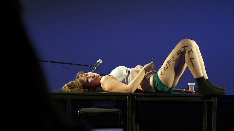 A performer lies across a table under blue stage light, with the word INFERTIL written along one leg.