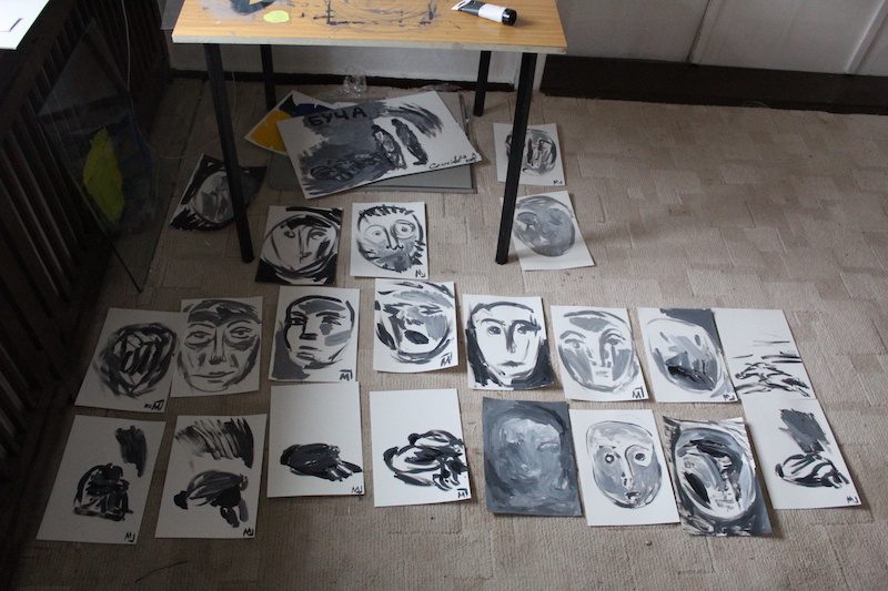 Dozens of black-and-white painted portraits on paper laid out across a studio floor around a work table.