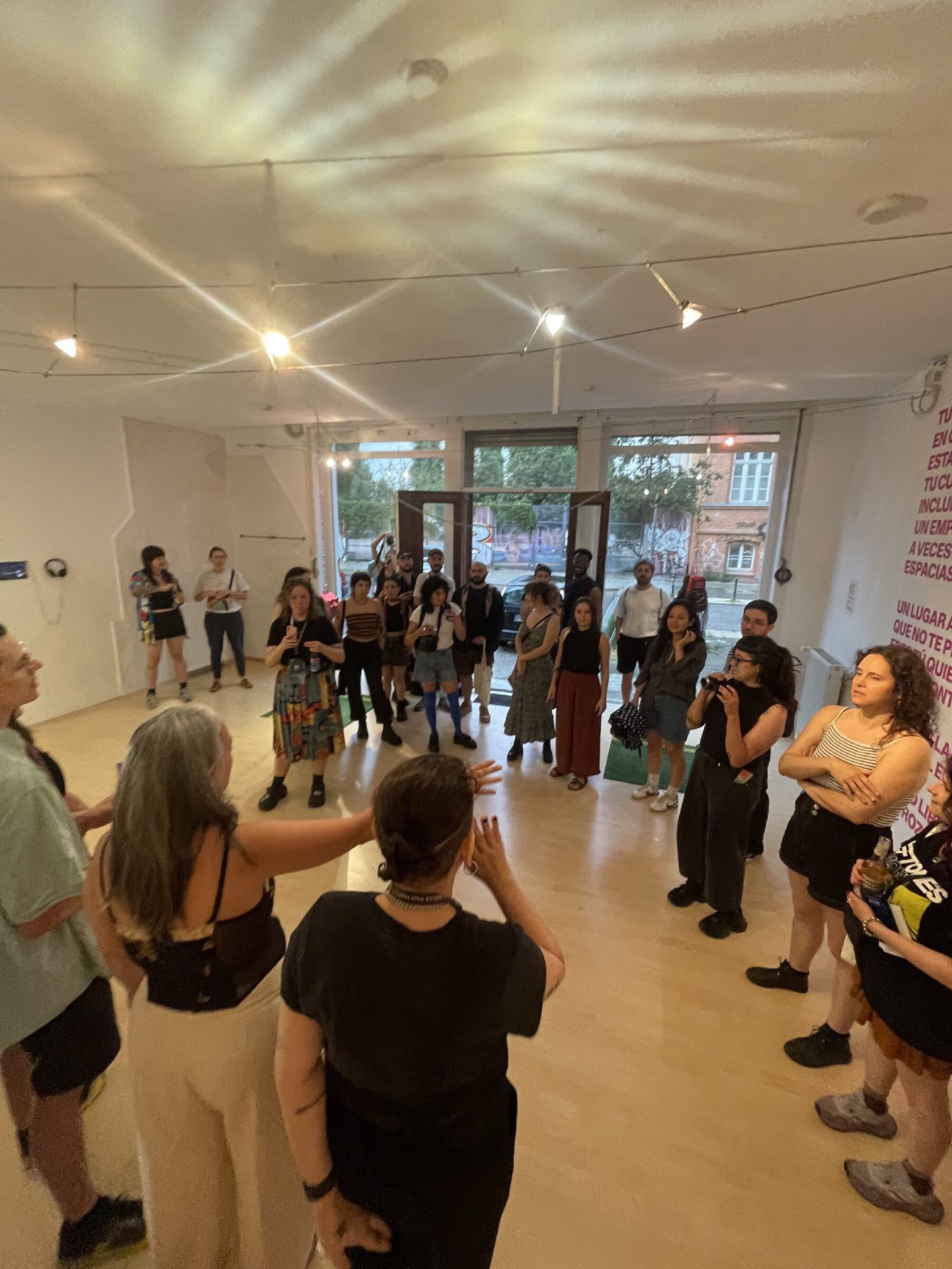 A large group of visitors gathered in the white-walled gallery at Bardo Projektraum, listening to a speaker in the foreground, with a wall text piece in pink lettering and string lights overhead.