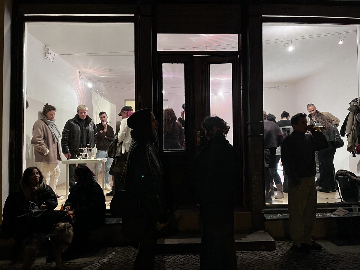 People spill onto the pavement outside Bardo Projektraum at night, with a lit exhibition visible through the large shopfront windows.