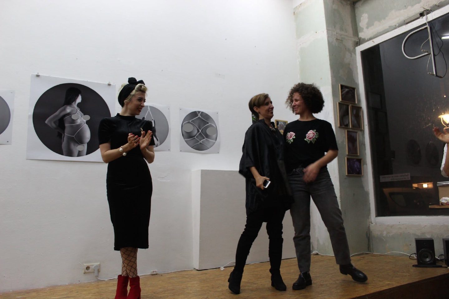 Three people stand and applaud in a gallery beside a row of circular black-and-white photographic works hung on a white wall, with warm lamplight from a window at the right.