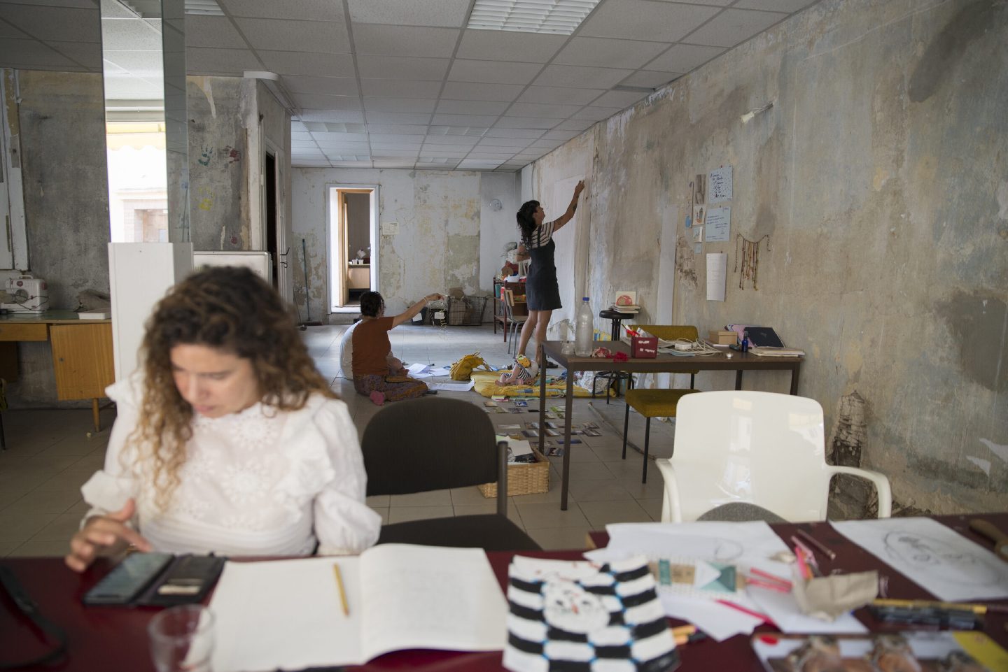 Artists at work in a large residency studio with peeling plaster walls: one writes at a table, another paints at the wall, and a child plays on the floor nearby.