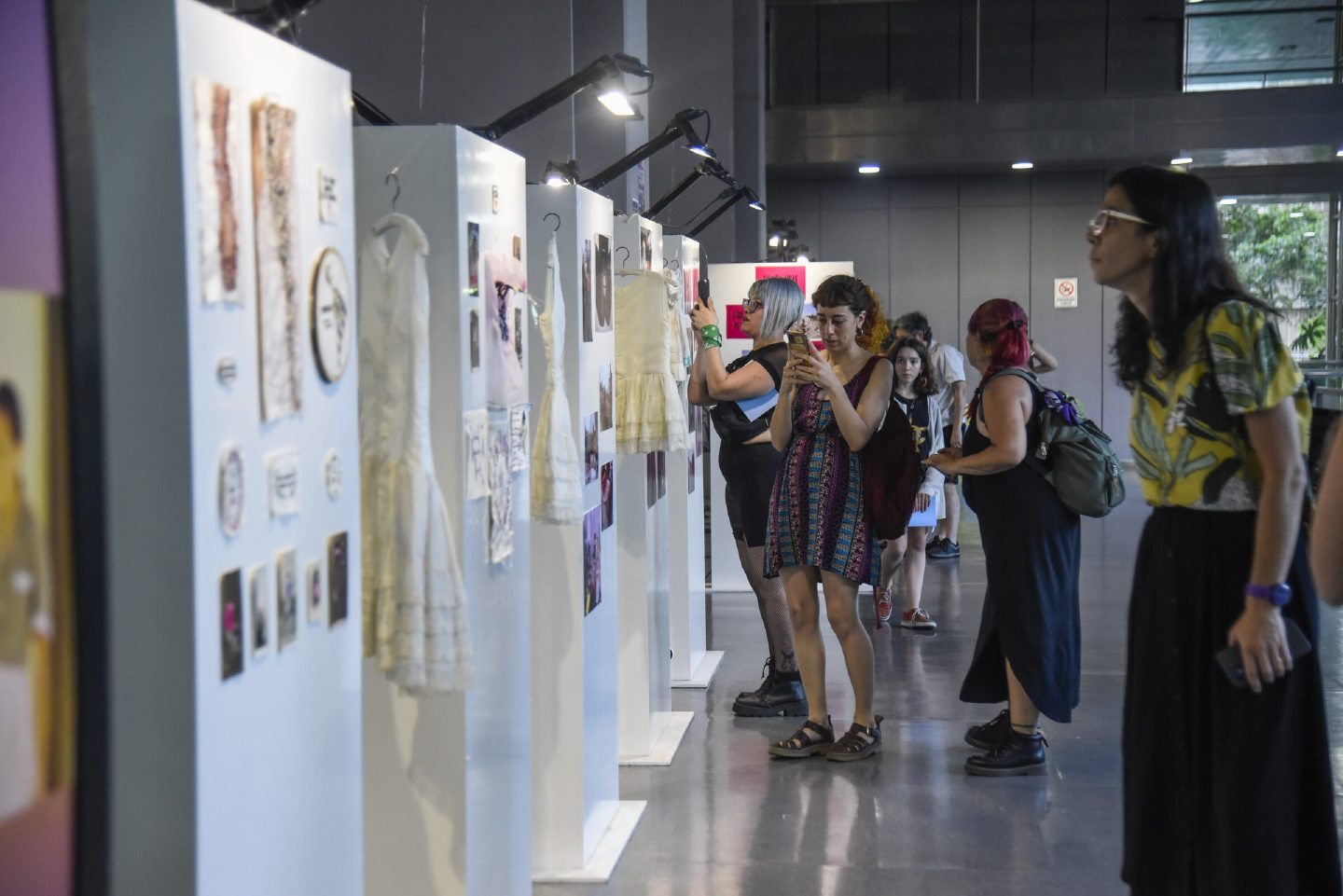 Visitors walk between white plinths hung with embroidered textiles, dresses and small framed works in a large hall.