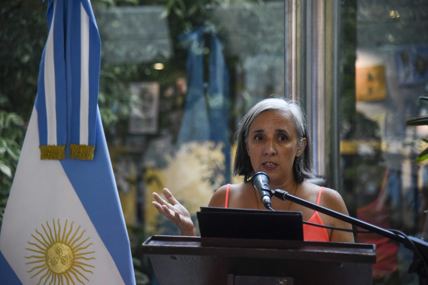 A speaker addresses an audience from a wooden lectern beside an Argentine flag.