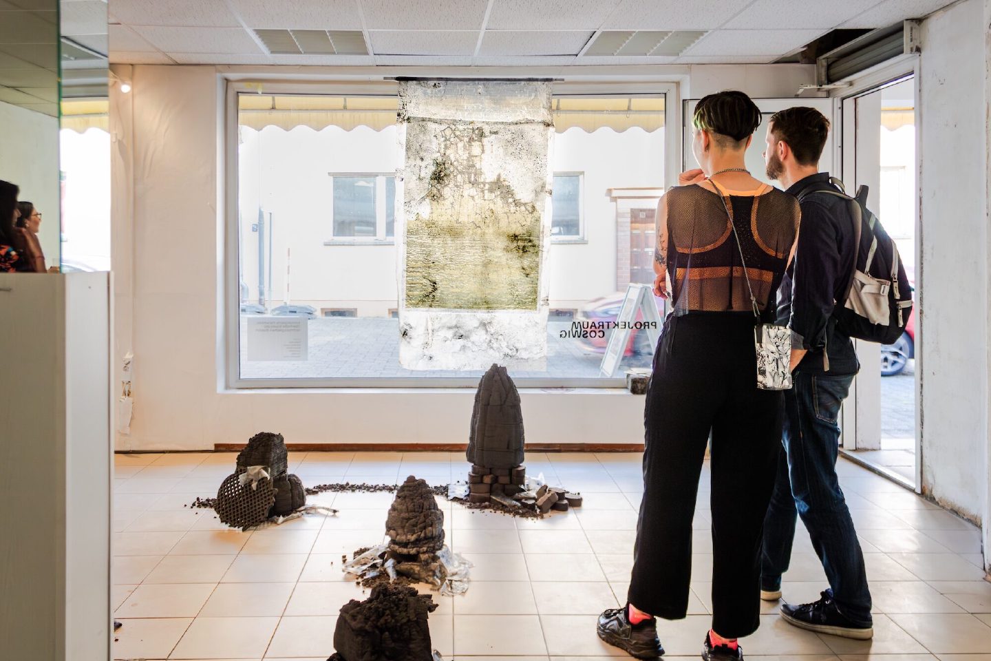 Two visitors look at a weathered hanging textile in a shopfront gallery, with small charred sculptural forms arranged across the tiled floor.