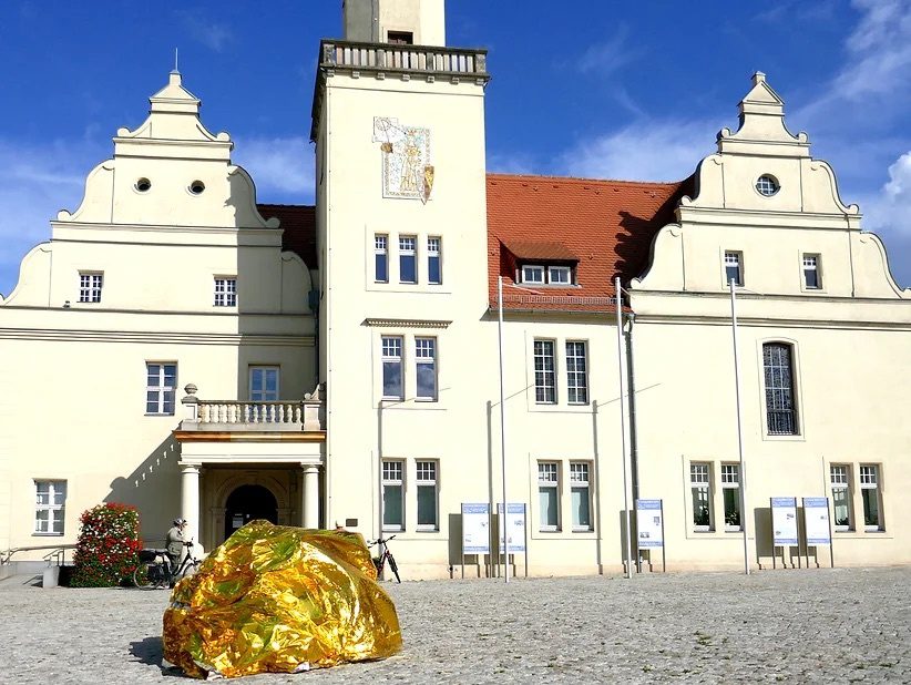 A gold foil-wrapped form rests on the cobbled forecourt of the white gabled Schloss Coswig under a blue sky.
