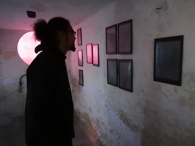 A visitor looks at framed works hung on a bare plastered cellar wall, lit by a large pink disc of light.