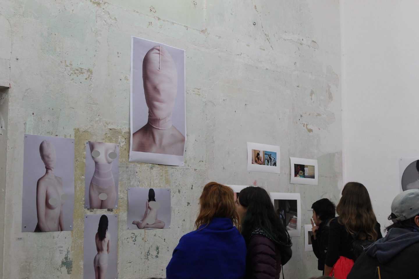 Visitors look at photographic works pinned to a bare plastered wall, among them large images of a figure with the head and face wrapped in fabric.