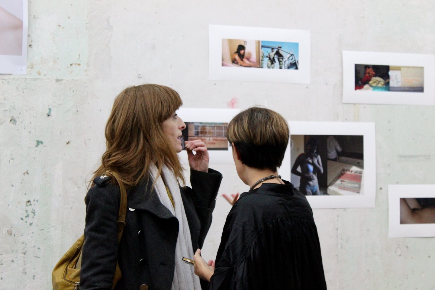 Two visitors talk in front of a wall of unframed colour photographs mounted on a rough plastered surface.