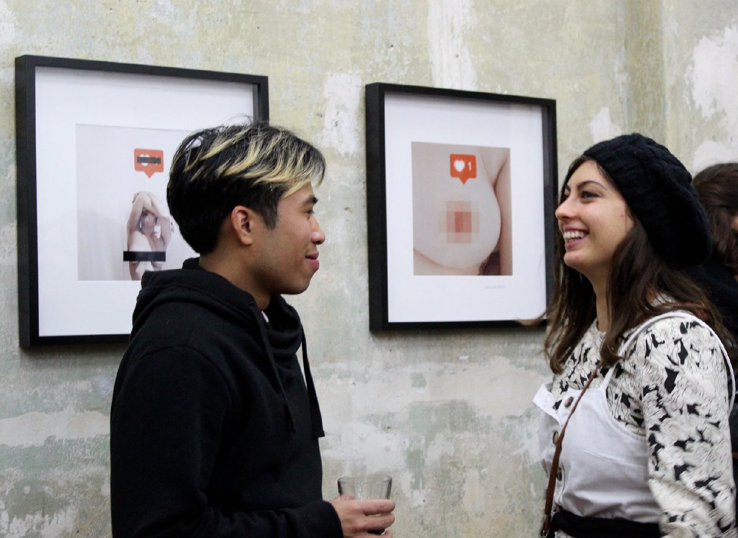 Two visitors laugh beside framed works in which a breast is covered by a pixellated block and a social-media “like” icon.