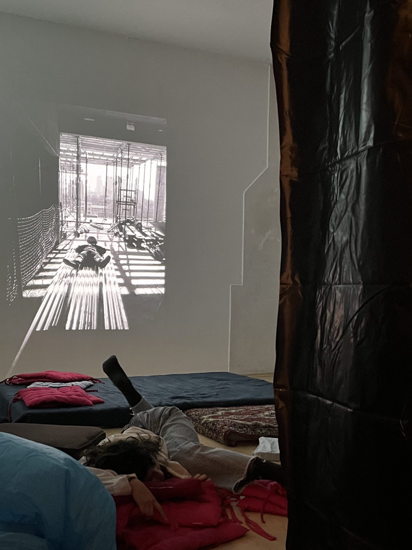 A darkened room lined with black tarpaulin where visitors rest on mattresses beneath a projected film of construction workers.