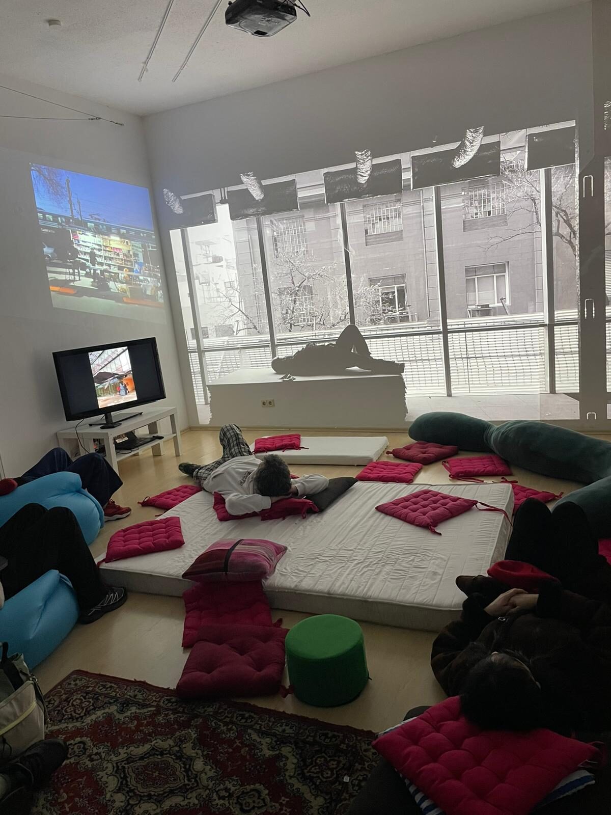 A light-filled room where visitors lie on a low mattress and floor cushions facing a projection, with photographs strung across the windows.