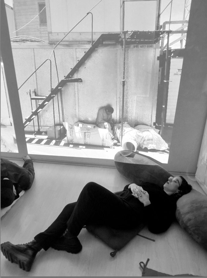 Black and white photograph: a person reclines on a beanbag indoors while, through the window, a worker sits resting among stacked cement sacks on a building site.