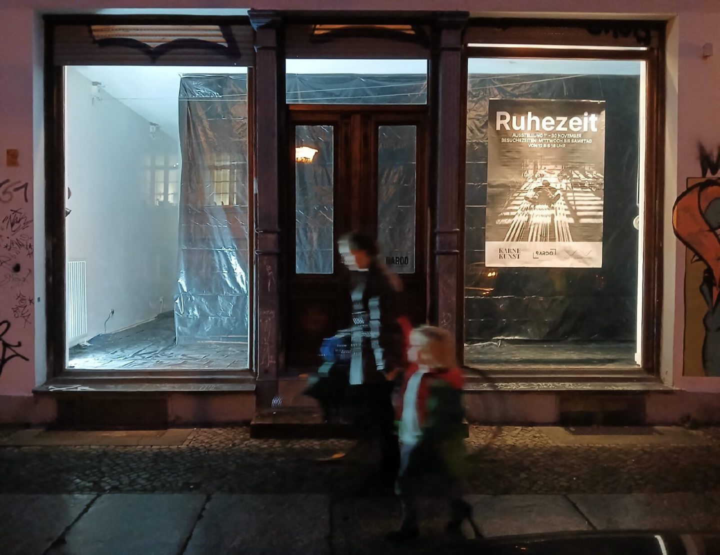 The lit shopfront of Bardo Projektraum at night with the Ruhezeit exhibition poster in the window as a woman and child walk past.