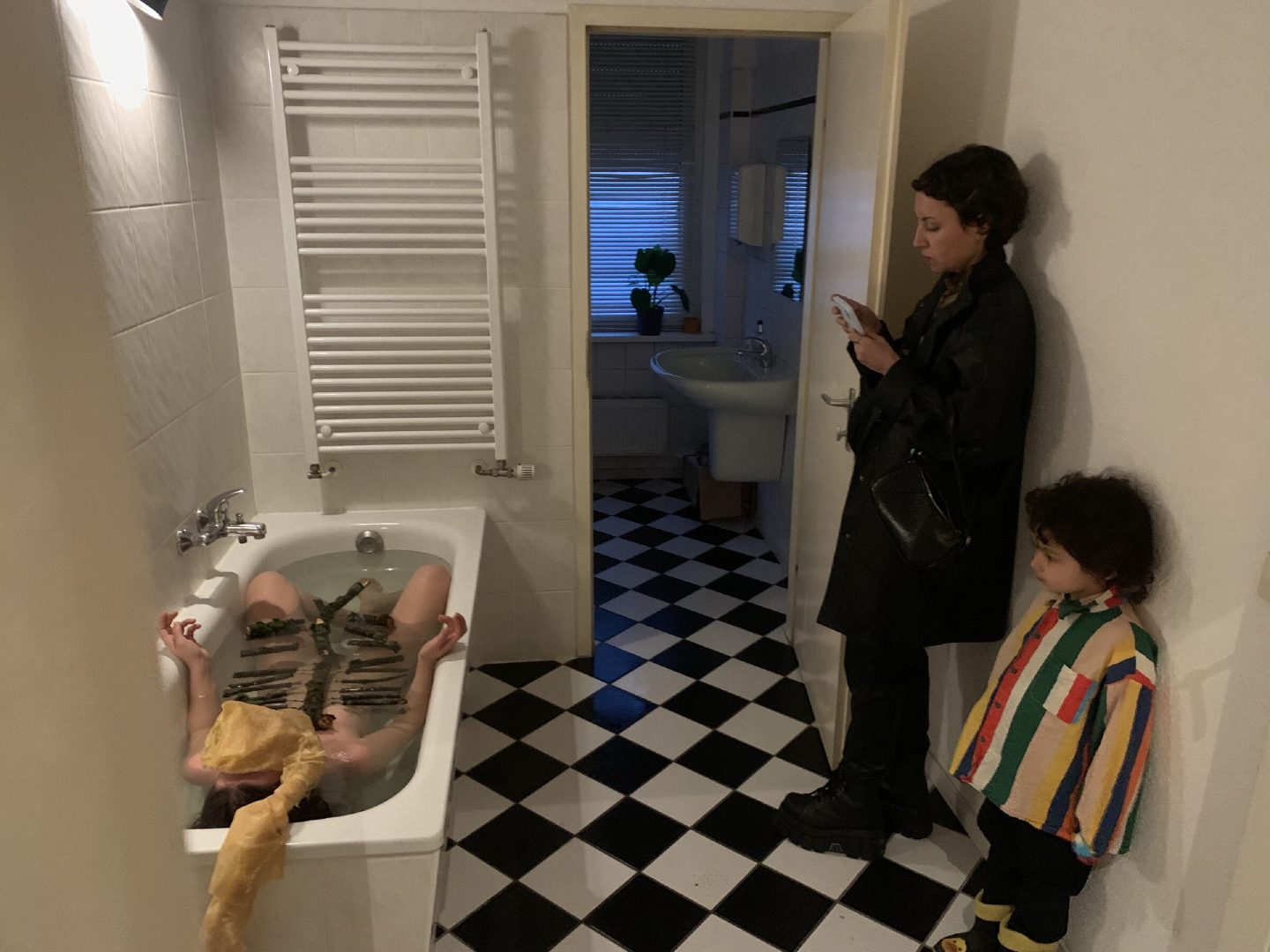 Visitors, one of them a child, watch a performer lying in a bathtub laid with branches.